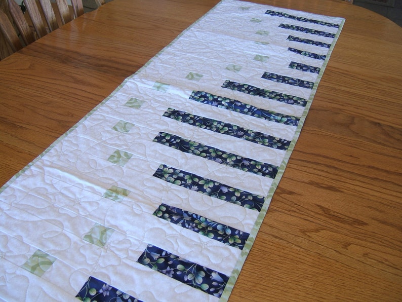 Pode incluir: Um corredor de mesa branco acolchoado com tiras de tecido com padr&atilde;o floral verde e azul. O corredor de mesa tem um design acolchoado com um padr&atilde;o floral.