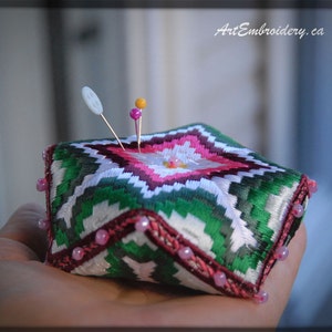 May include: A green, white, and pink pincushion with a geometric pattern and pink beads around the edges. The pincushion is filled with pins and has a white button on top.