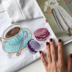 Peut inclure: Un motif brodé sur un t-shirt blanc représentant une tasse de café avec une soucoupe, une cuillère et deux macarons. Les macarons sont rose et violet.