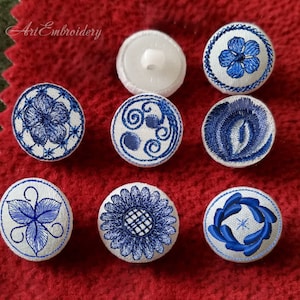 Può includere: Otto bottoni in tessuto bianco con disegni ricamati in blu. I disegni includono un fiore, un vortice, una foglia, un girasole e una ghirlanda.