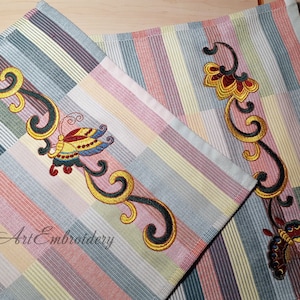 Puede incluir: Dos toallas de té bordadas con un patrón a rayas en tonos de rosa, amarillo, azul y gris. Las toallas presentan mariposas y diseños florales bordados en hilo dorado y negro. El texto "Art Embroidery" es visible en una de las toallas.