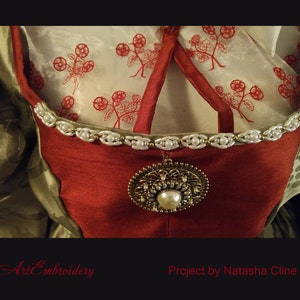 May include: Close-up of a red and green dress with a white pearl trim and a gold circular brooch. The dress has a white fabric with red floral embroidery underneath.
