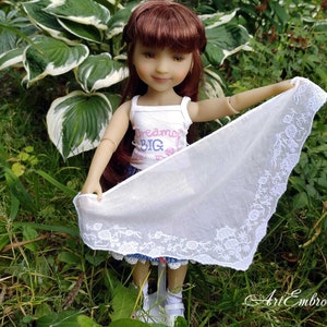 Puede incluir: Una muñeca con un top blanco sin mangas con el texto "Dream Big" en letras rosa y azul, una falda azul y blanca y una bufanda de encaje blanco. La muñeca sostiene la bufanda en sus manos y está de pie en un área cubierta de hierba.