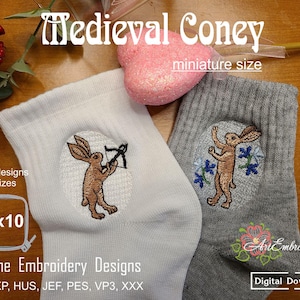 Puede incluir: Calcetines blancos y grises con diseños de conejos bordados. Un conejo sostiene un arco y una flecha, el otro está de pie con los brazos levantados. El texto "Medieval Coney" y "miniature size" son visibles. El diseño es 15.24 x 25.4 cm.