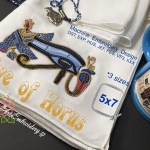 Puede incluir: Tela blanca bordada con el diseño del Ojo de Horus en azul, dorado y rojo. El texto "Eye of Horus" es dorado. El diseño está etiquetado como "Machine Embroidery Design" e incluye información de tamaño "5x7".