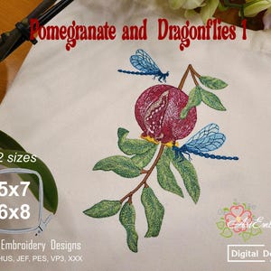 Puede incluir: Diseño bordado con una granada y dos libélulas. La granada es de color rojo intenso con hojas verdes y tallos marrones. Dos libélulas azules vuelan alrededor de la fruta. El texto dice "Pomegranate and Dragonflies 1".