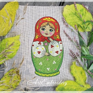 May include: Embroidered design of a traditional Russian nesting doll, or Matryoshka doll, with a red headscarf, green dress, and floral details. The doll is standing on a beige fabric background.