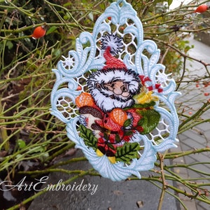 May include: A white lace Christmas ornament with a blue border featuring a Santa Claus design with a red hat, white beard, and holding a red and green gift. The ornament is hanging on a branch with green leaves and red berries.