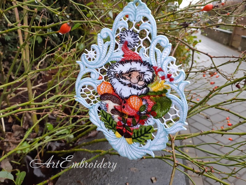 May include: A white lace Christmas ornament with a blue border featuring a Santa Claus design with a red hat, white beard, and holding a red and green gift. The ornament is hanging on a branch with green leaves and red berries.