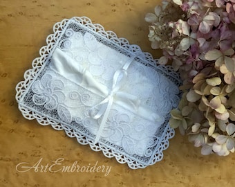 May include: White lace ring pillow with a satin ribbon tied around it. The pillow is on a brown wooden surface with dried flowers in the background.