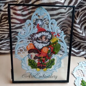 May include: A framed embroidery design featuring a blue and white lace pattern with a Santa Claus figure holding a bag of gifts. The embroidery is titled "Art Embroidery".