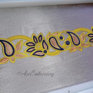 Può includere: Primo piano di un ricamo oro e blu su un tessuto grigio chiaro. Il disegno presenta forme a paisley e motivi floreali. Il testo "Art Embroidery" è visibile nella parte inferiore dell'immagine.