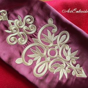 Peut inclure: Gros plan sur un tissu bordeaux avec des broderies dorées. La broderie présente un motif floral avec des volutes et des feuilles complexes. Le texte "Art Embroidery" est visible dans le coin supérieur droit de l'image.