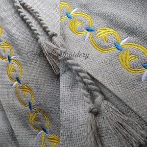 Puede incluir: Primer plano de una tela de lino beige con un diseño bordado en amarillo y azul. La tela tiene un borla de cuerda.