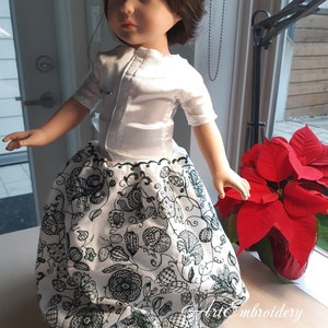 May include: A white doll wearing a white blouse with a green embroidered skirt. The skirt has a floral pattern with green leaves and flowers. The doll has brown hair and blue eyes.