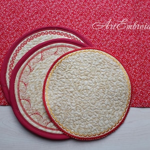 May include: Three round quilted placemats with embroidered designs. The largest placemat is cream with a gold embroidered border and a swirling pattern. The two smaller placemats are red with gold embroidered borders and a floral design. The placemats are on a white surface.