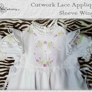 May include: White baby dress with a cross and floral embroidery. The dress has lace trim and a cutwork lace applique on the sleeves. The text "Cutwork Lace Applique Sleeve Wing 1" is at the top of the image.