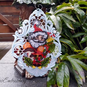 May include: A white, blue, and green embroidered Christmas ornament featuring a Santa Claus figure holding a bag of gifts and mushrooms. The ornament is hanging from a wire loop.
