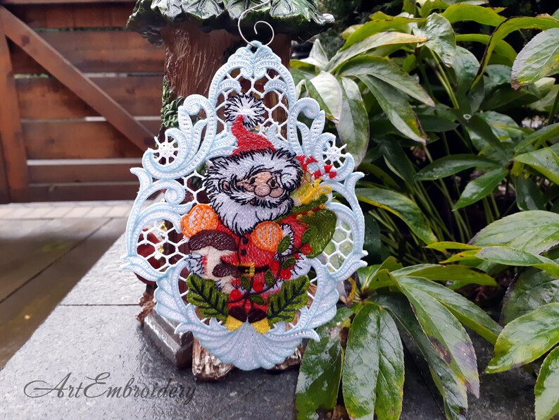 May include: A white, blue, and green embroidered Christmas ornament featuring a Santa Claus figure holding a bag of gifts and mushrooms. The ornament is hanging from a wire loop.