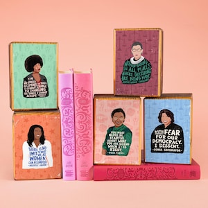 May include: A collection of decorative wooden blocks and books. The blocks feature portraits of notable women with inspirational quotes. The books are pink with decorative patterns. The quotes include Michelle Obama, Rosa Parks, and Sonia Sotomayor.