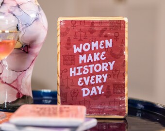 Könnte beinhalten: Ein Holzschild mit rotem Hintergrund und weißem Text, der "Frauen machen Geschichte jeden Tag" lautet. Das Schild ist mit kleinen weißen feministischen Symbolen verziert.