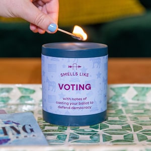 May include: A lit candle in a dark blue container with the words "Smells Like Voting" and "with notes of casting your ballot to defend democracy." The candle is on a decorative tray.