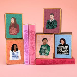 Peut inclure: Quatre blocs rectangulaires en bois illustrés de portraits de personnages historiques avec des citations, empilés avec deux livres roses et un livre rouge. Les blocs présentent Gloria Steinem, Ruth Bader Ginsburg, Rosa Parks et Sonia Sotomayor.