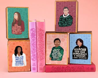 Könnte beinhalten: Vier rechteckige Holzblöcke mit Illustrationen historischer Persönlichkeiten und Zitaten, gestapelt neben zwei rosa und einem roten Buch. Die Blöcke zeigen Porträts von Gloria Steinem, Ruth Bader Ginsburg, Rosa Parks und Sonia Sotomayor.