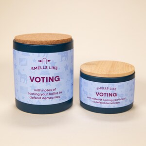 May include: Two cylindrical candles with lids. The larger candle has a cork lid, and the smaller one has a wooden lid. Both candles are dark blue with a light blue band featuring stars, flags, and the text "SMELLS LIKE VOTING".