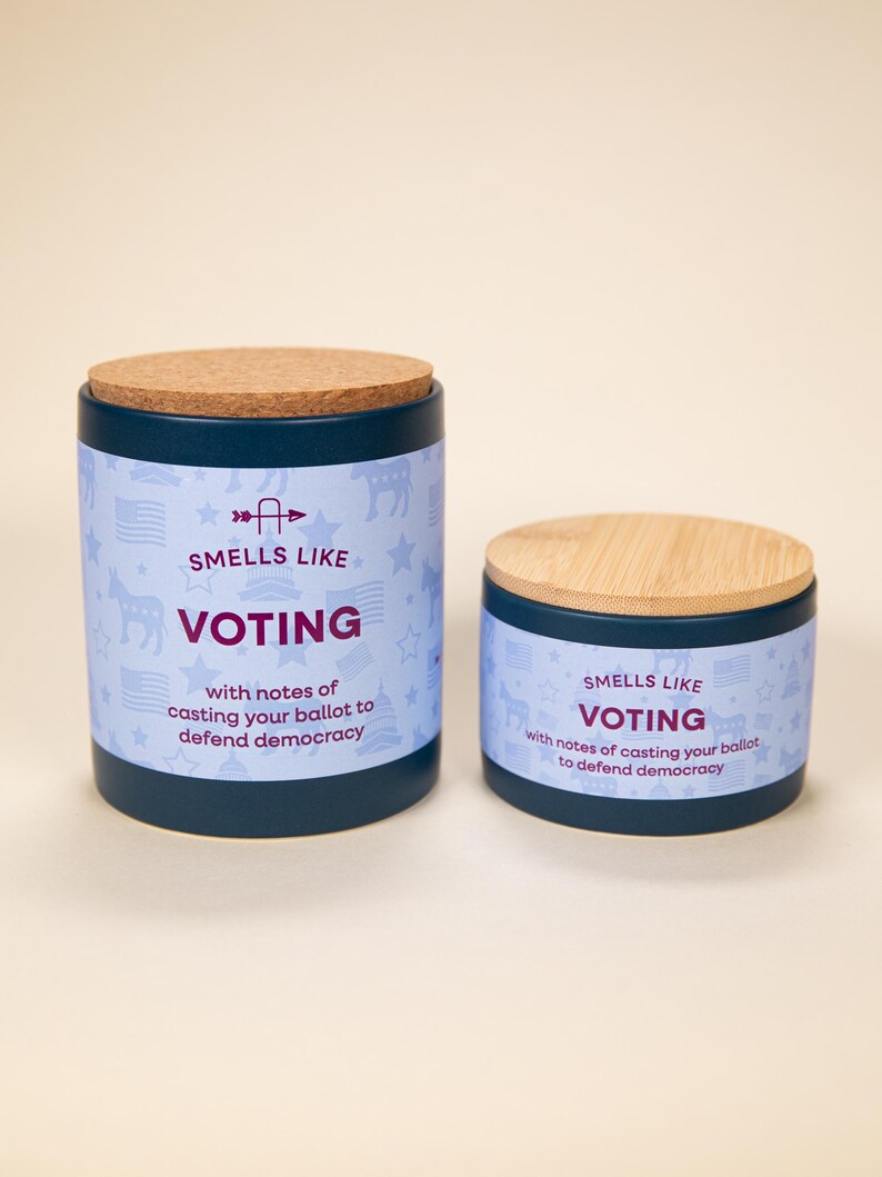 May include: Two cylindrical candles with lids. The larger candle has a cork lid, and the smaller one has a wooden lid. Both candles are dark blue with a light blue band featuring stars, flags, and the text "SMELLS LIKE VOTING".
