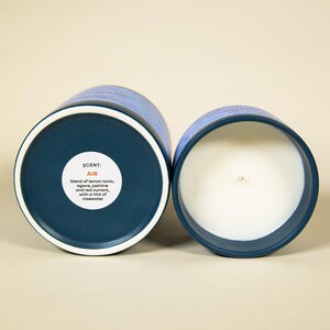 May include: Two blue and white scented candles. One candle is open, revealing a white wax interior and a wick. The other candle is closed, showing the bottom with the scent description: "AIR" with notes of lemon tonic, agave, jasmine, red currant, and rosewater.