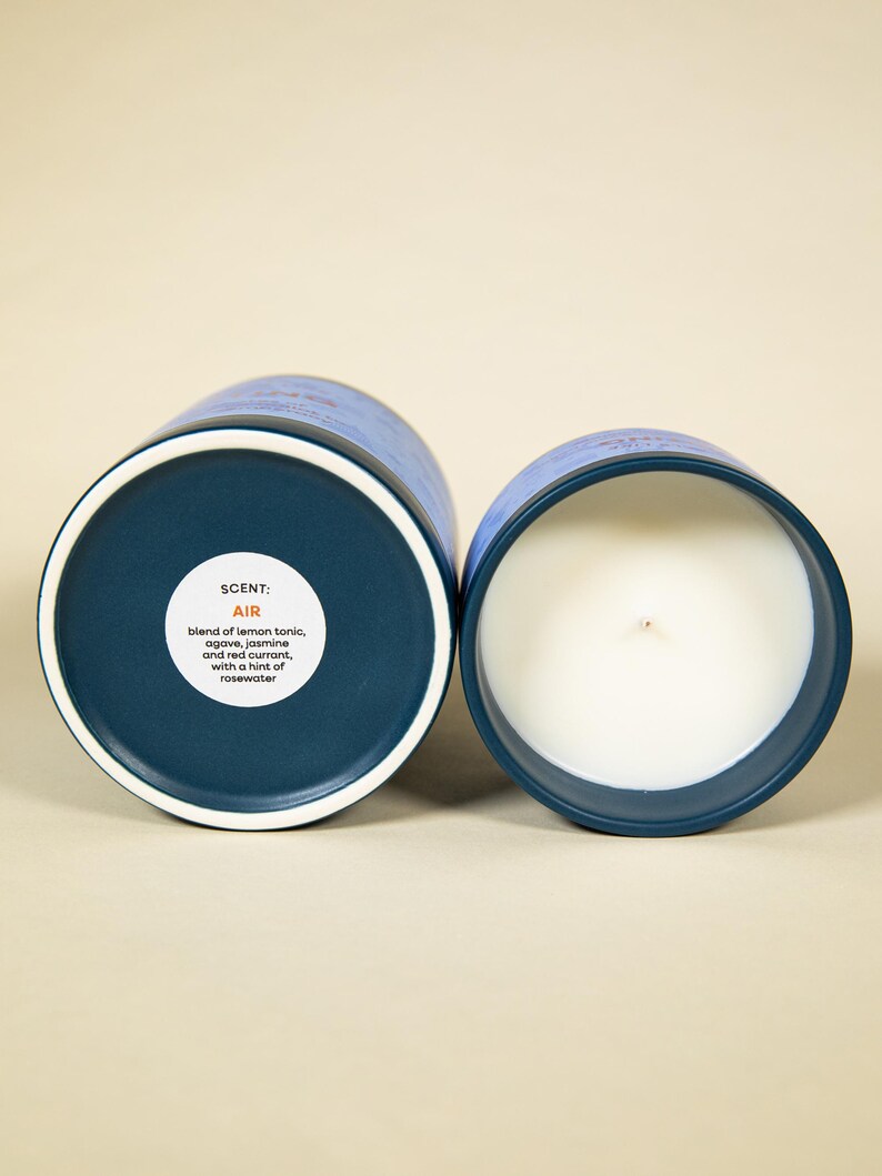 May include: Two blue and white scented candles. One candle is open, revealing a white wax interior and a wick. The other candle is closed, showing the bottom with the scent description: "AIR" with notes of lemon tonic, agave, jasmine, red currant, and rosewater.