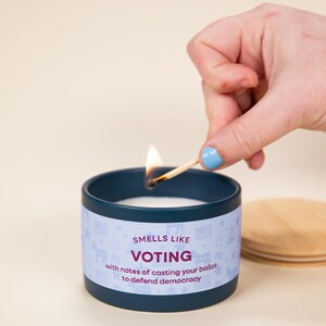 May include: A navy blue candle in a tin with the text "Smells Like Voting" and a light blue label with stars, flags, and a unicorn. A lit match is held above the candle. A wooden lid is next to the candle.