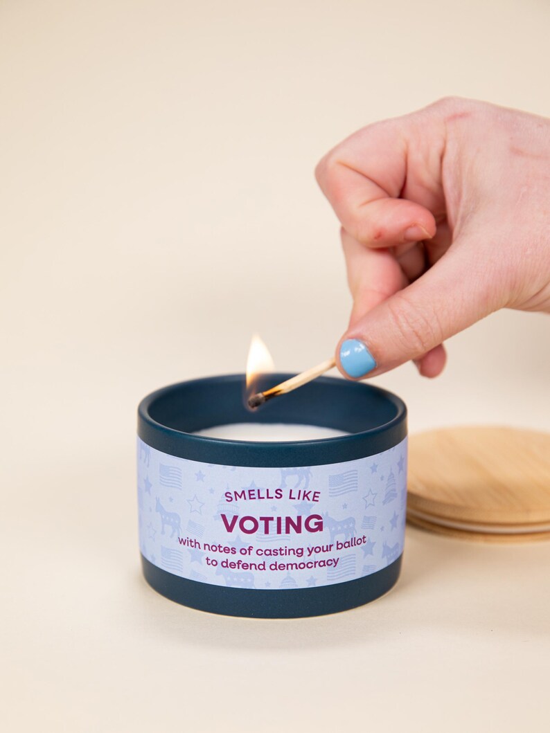 May include: A navy blue candle in a tin with the text "Smells Like Voting" and a light blue label with stars, flags, and a unicorn. A lit match is held above the candle. A wooden lid is next to the candle.