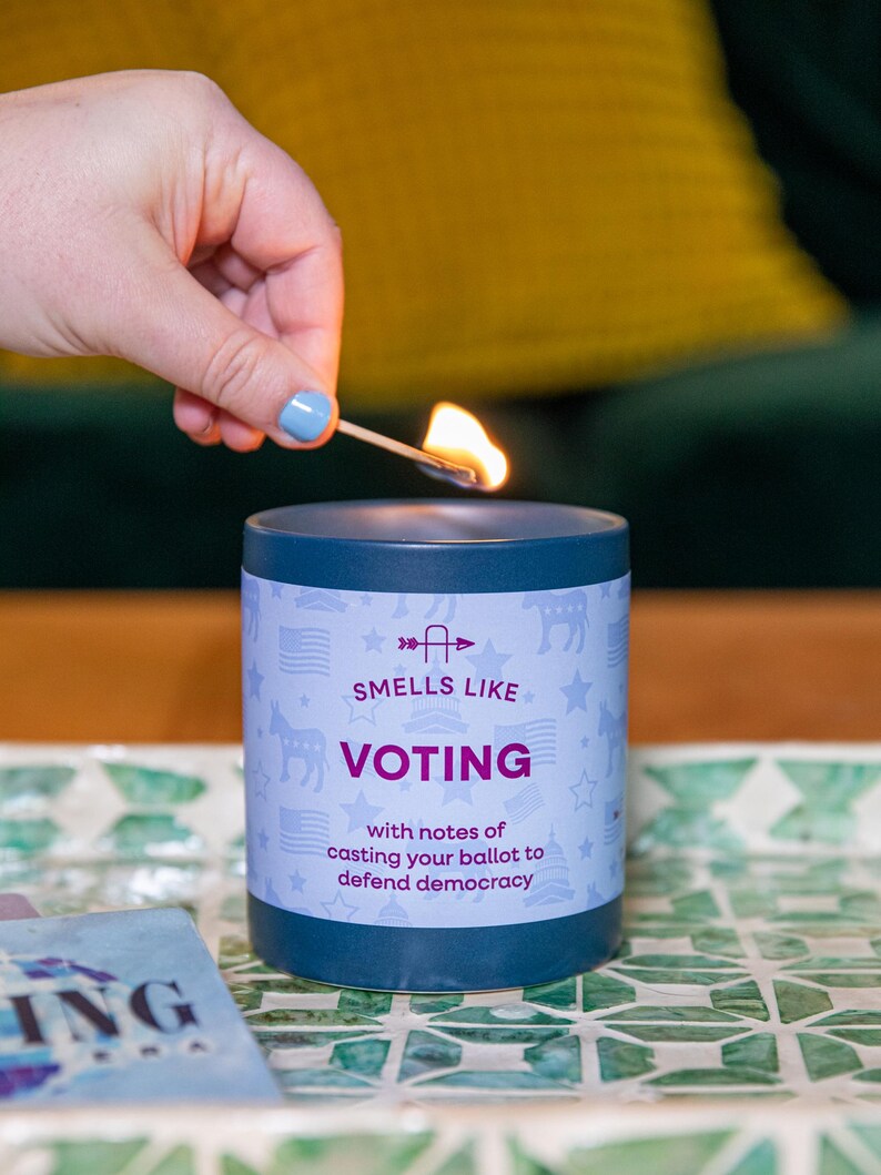 May include: A lit candle in a dark blue container with the words "Smells Like Voting" and "with notes of casting your ballot to defend democracy." The candle is on a decorative tray.