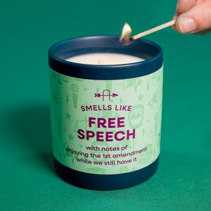 Puede incluir: Una vela encendida en un recipiente de cerámica azul oscuro. La vela tiene una etiqueta verde claro con las palabras "SMELLS LIKE FREE SPEECH" y otro texto. Se está utilizando una cerilla para encender la vela.