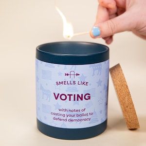 May include: A navy blue candle with the text "Smells Like Voting" and a light blue background with patriotic symbols. A match is lit above the candle. The candle has a wooden lid.