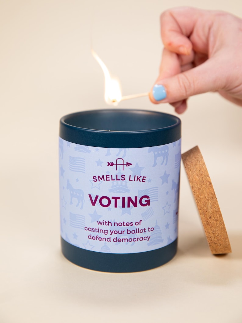 May include: A navy blue candle with the text "Smells Like Voting" and a light blue background with patriotic symbols. A match is lit above the candle. The candle has a wooden lid.