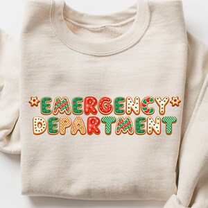 May include: A beige sweatshirt with the words "EMERGENCY DEPARTMENT" in a gingerbread cookie font. The letters are red, green, and white with decorative icing. The sweatshirt is folded, with the sleeves visible.