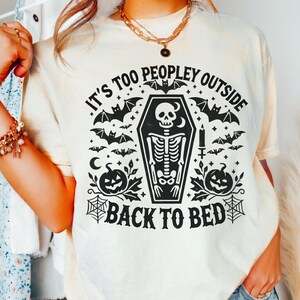 May include: A white t-shirt with a black graphic print of a skeleton in a coffin surrounded by bats, pumpkins, and the text "It's too peopley outside back to bed."
