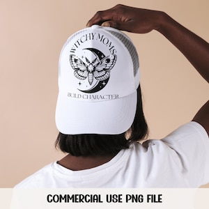 K&ouml;nnte beinhalten: Wei&szlig;e Trucker-Kappe mit einem schwarz-wei&szlig;en Motiv aus einem Nachtfalter und einem Halbmond. Der Text "Witchy Moms Build Character" ist auf der Vorderseite der Kappe aufgedruckt.