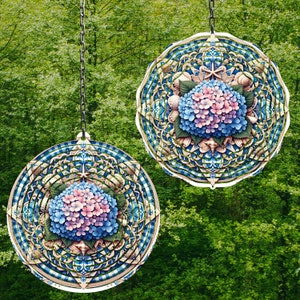 May include: Two round, decorative hanging ornaments with a floral and mandala design. Each ornament features a central hydrangea flower in shades of pink and blue, surrounded by intricate patterns in blue, white, and beige. The ornaments hang from a chain against a green background.