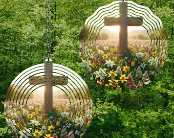 May include: Two metal wind spinners with a cross design and a field of flowers in the center. The spinners are hanging from a tree branch and are surrounded by green leaves.