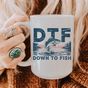 May include: White ceramic mug with the text "DTF" above a graphic of a fish jumping out of the water. The words "DOWN TO FISH" are below the graphic. The mug is held by a person.