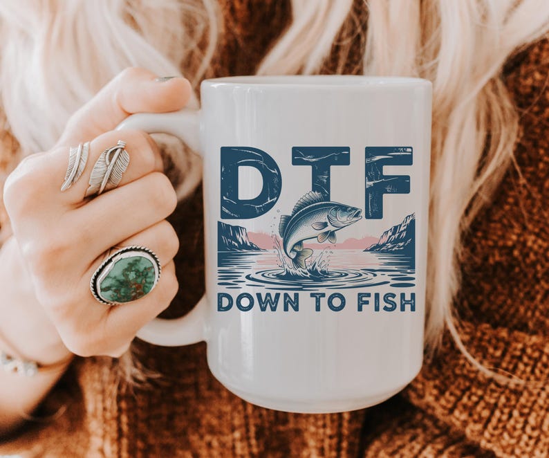 May include: White ceramic mug with the text "DTF" above a graphic of a fish jumping out of the water. The words "DOWN TO FISH" are below the graphic. The mug is held by a person.