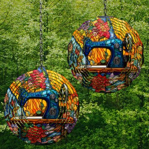 May include: Two round stained glass ornaments with a vintage sewing machine design. The ornaments are hanging from chains against a green leafy background. The sewing machines are blue and yellow with red and orange accents.
