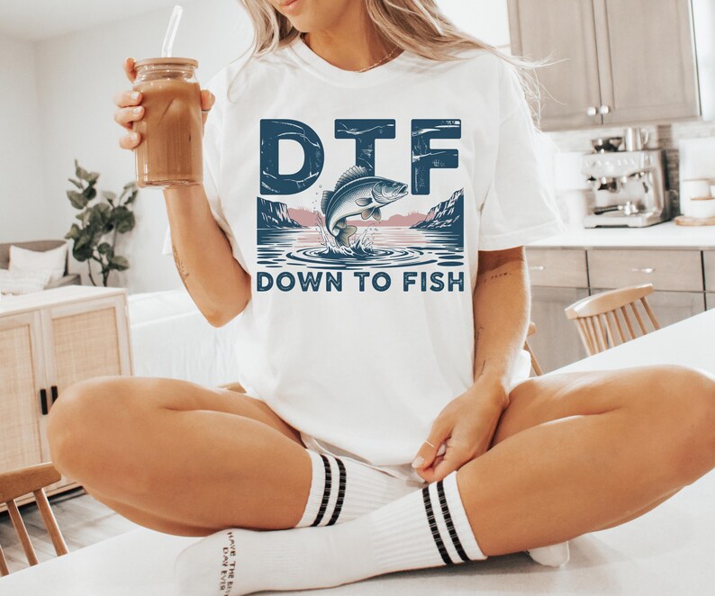 May include: White t-shirt with a graphic of a fish jumping out of water, with the text "DTF DOWN TO FISH" in blue. The shirt is a casual style, suitable for everyday wear.