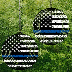 May include: Two black and white wind spinners with a blue line and a distressed look. The spinners are shaped like a circle and have a design that resembles a flag. The spinners are hanging from chains.