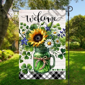 May include: A welcome garden flag with a white wood background and black and white checkered border. A mason jar filled with sunflowers, daisies, and clover is in the center of the flag. The jar has a green horseshoe tied around it with a brown ribbon.