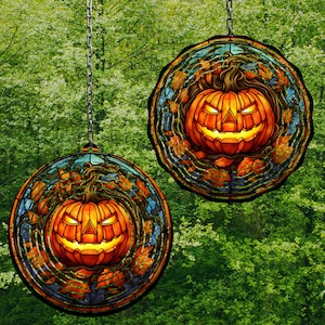 Op de afbeelding: Twee ronde glas-in-lood pompoen decoraties met een zwarte metalen ketting die van boven hangt. Elke pompoen is oranje met een zwarte steel en een gesneden jack-o'-lantern gezicht. De pompoenen zijn omringd door groene en bruine bladeren.